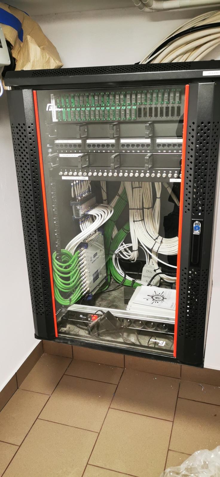 Szafa rack z okablowaniem strukturalnym: zielone i białe przewody uporządkowane wewnątrz czarnej szafy z pomarańczowymi akcentami, widok z przodu na tle beżowych płytek.