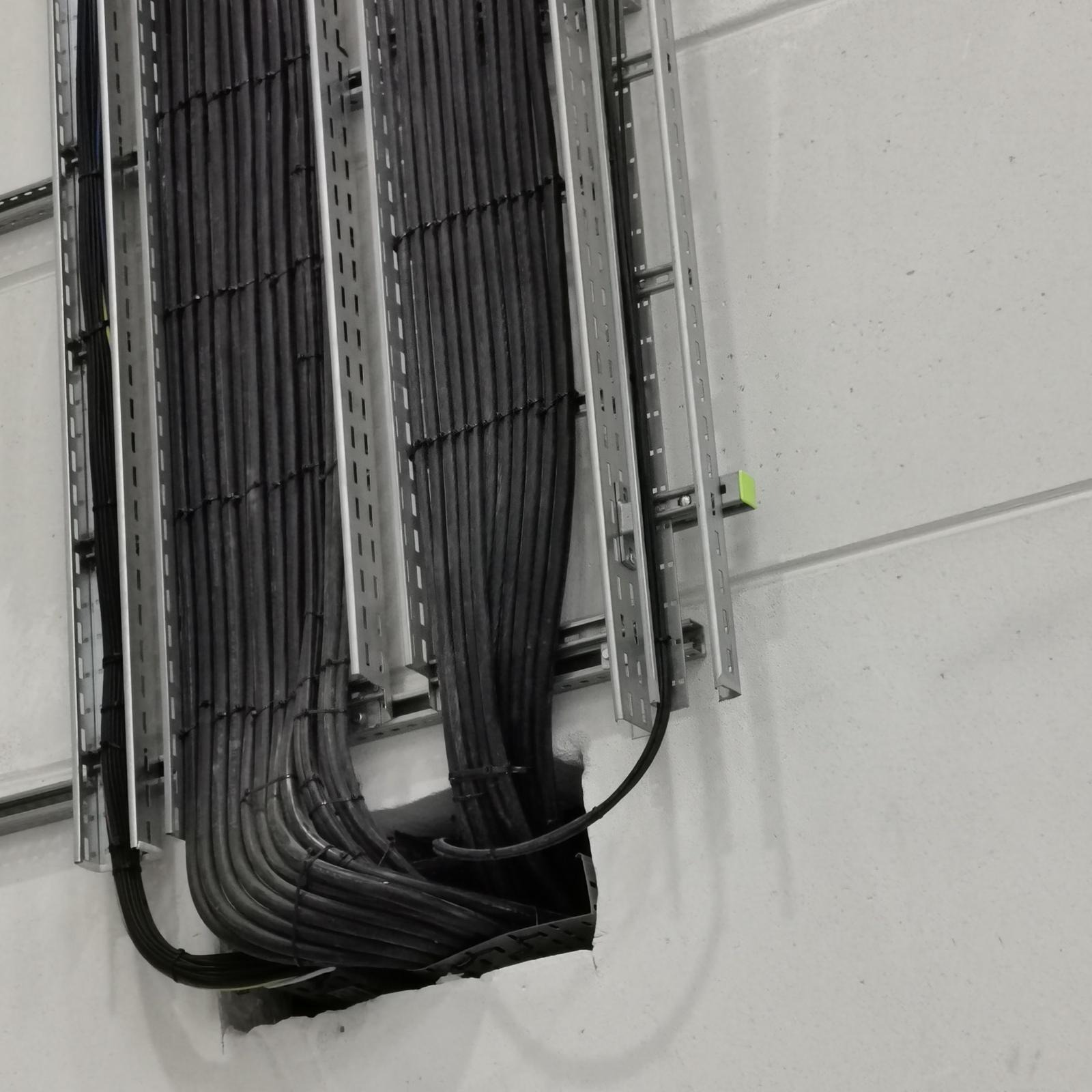 Kable elektryczne w metalowych korytkach przymocowanych do ściany. Widoczne prowadzenie przewodów przez otwór w ścianie, instalacja w szarym, surowym wnętrzu.