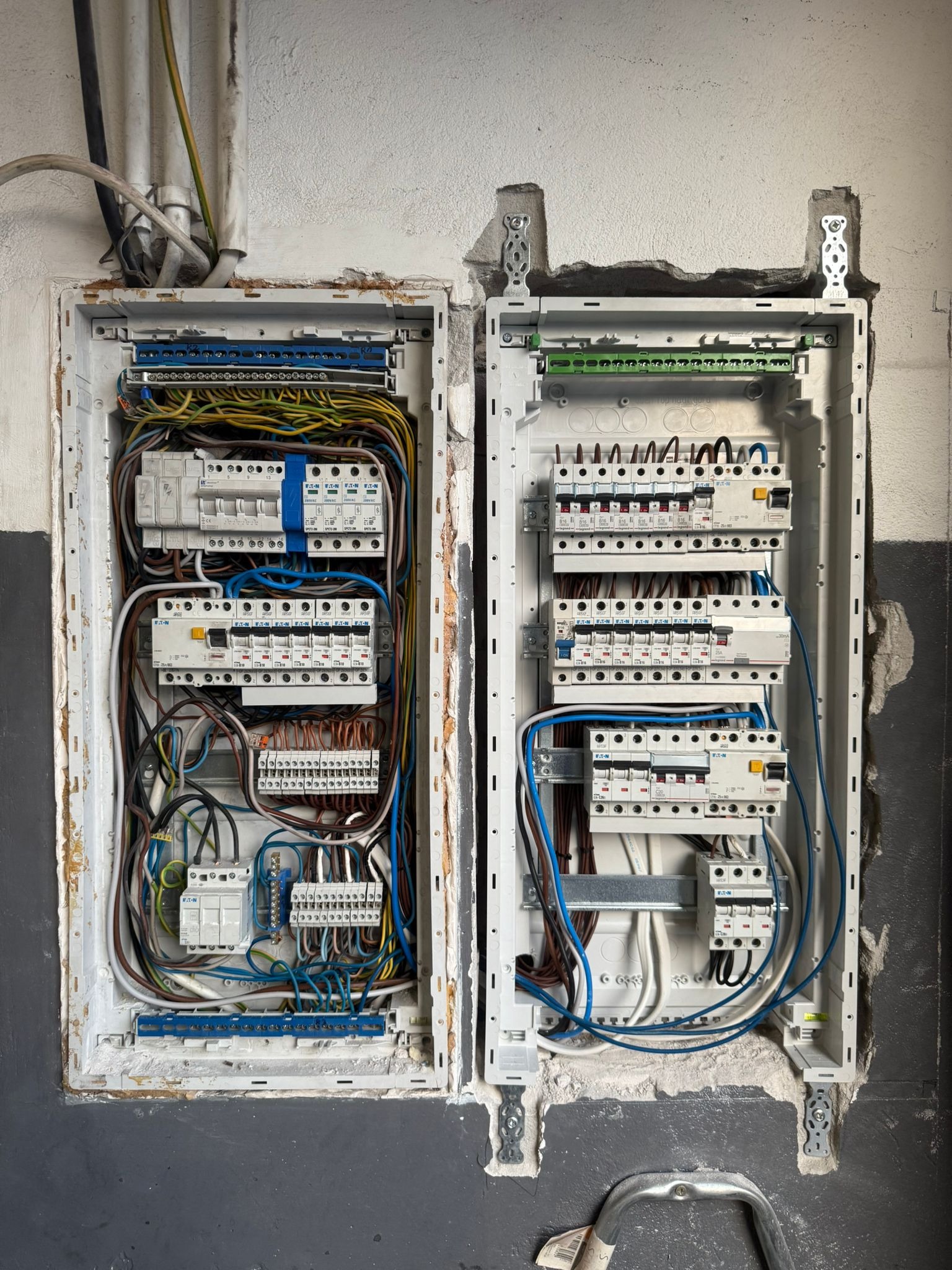 Dwa otwarte panele elektryczne z zainstalowanymi bezpiecznikami i okablowaniem, zamontowane w ścianie z surowym wykończeniem. Widoczne elementy montażowe i przewody.