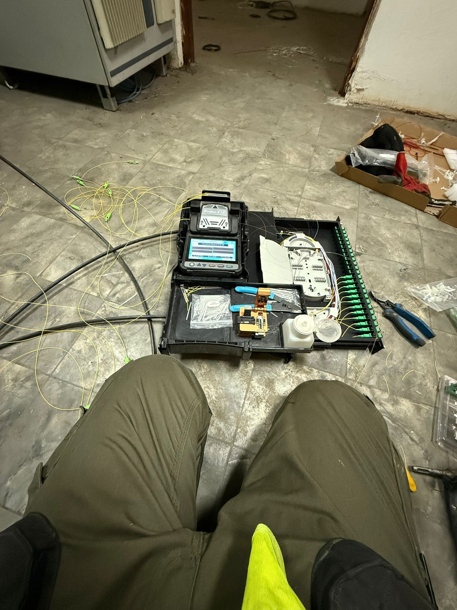 Otwarta skrzynka z narzędziami i światłowodami na podłodze. Widoczne złącza światłowodowe, spawarka światłowodowa i inne narzędzia. Praca w toku, instalacja telekomunikacyjna.