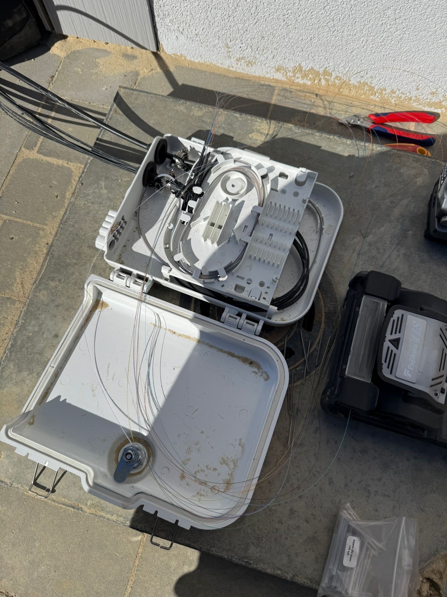 Otwarta skrzynka telekomunikacyjna z widocznymi światłowodami, narzędzia (obcinaczki) oraz spawarka światłowodowa Fujikura 41S na szarym tle kostki brukowej.