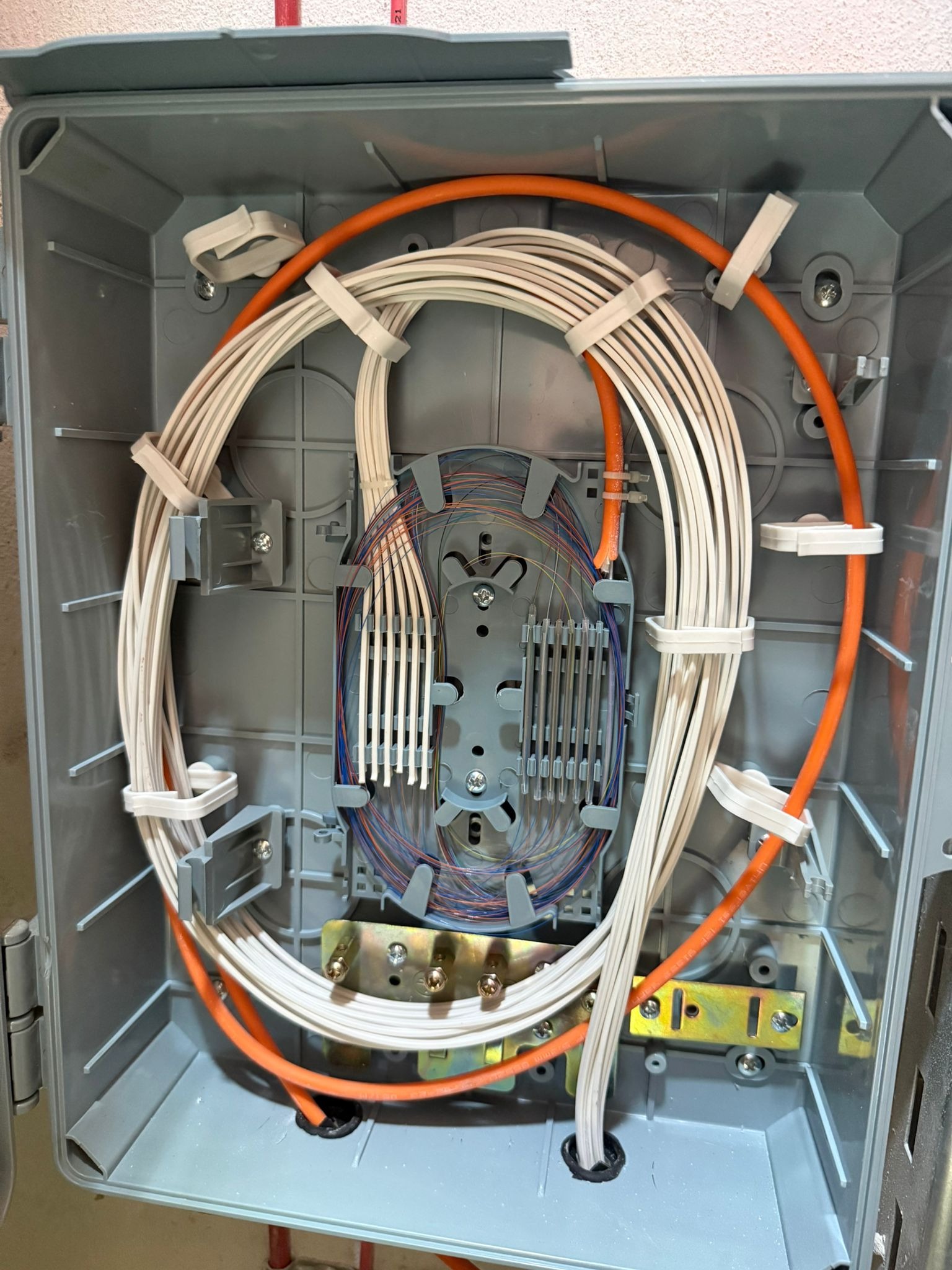 Otwarta szara skrzynka z uporządkowanymi kablami światłowodowymi, zabezpieczonymi białymi opaskami i pomarańczową rurką karbowaną. Widok z góry.