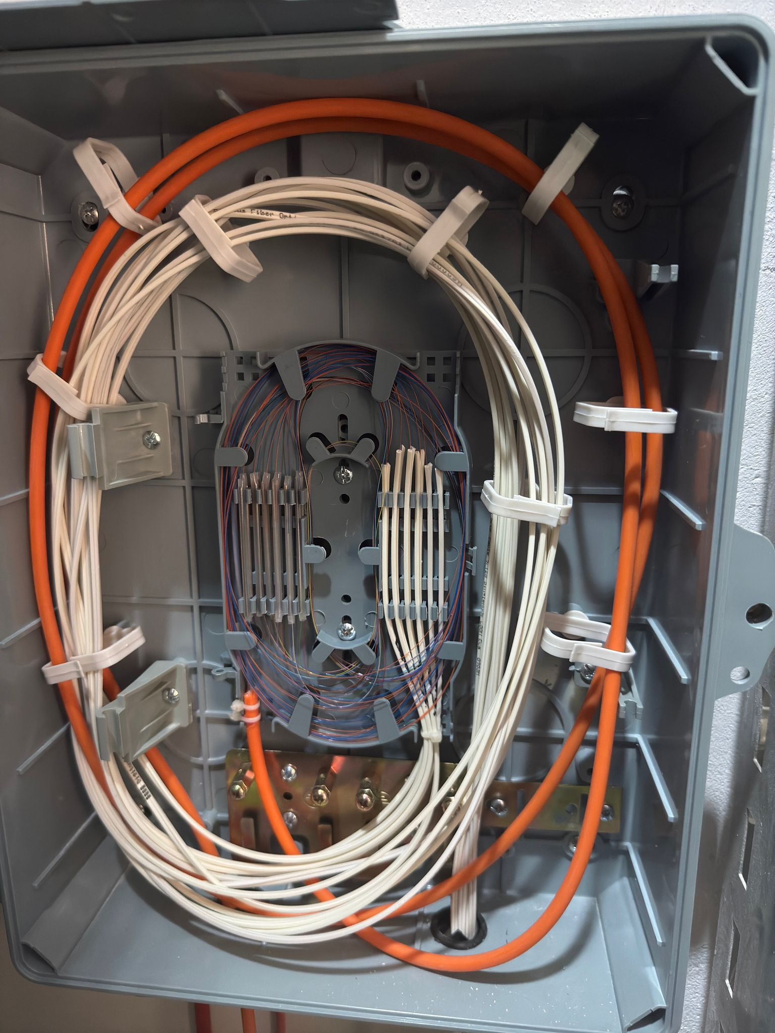 Szara skrzynka z uporządkowanymi kablami światłowodowymi (białe) i pomarańczowymi przewodami, zabezpieczonymi białymi opaskami, wewnątrz skrzynki.