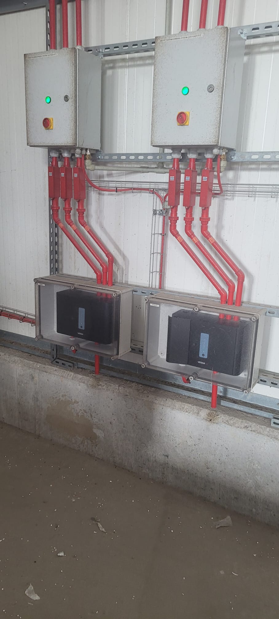 Dwa szare panele sterowania z zielonymi diodami i czerwonymi przyciskami awaryjnymi, połączone czerwonymi rurami z czujnikami w przezroczystych obudowach.