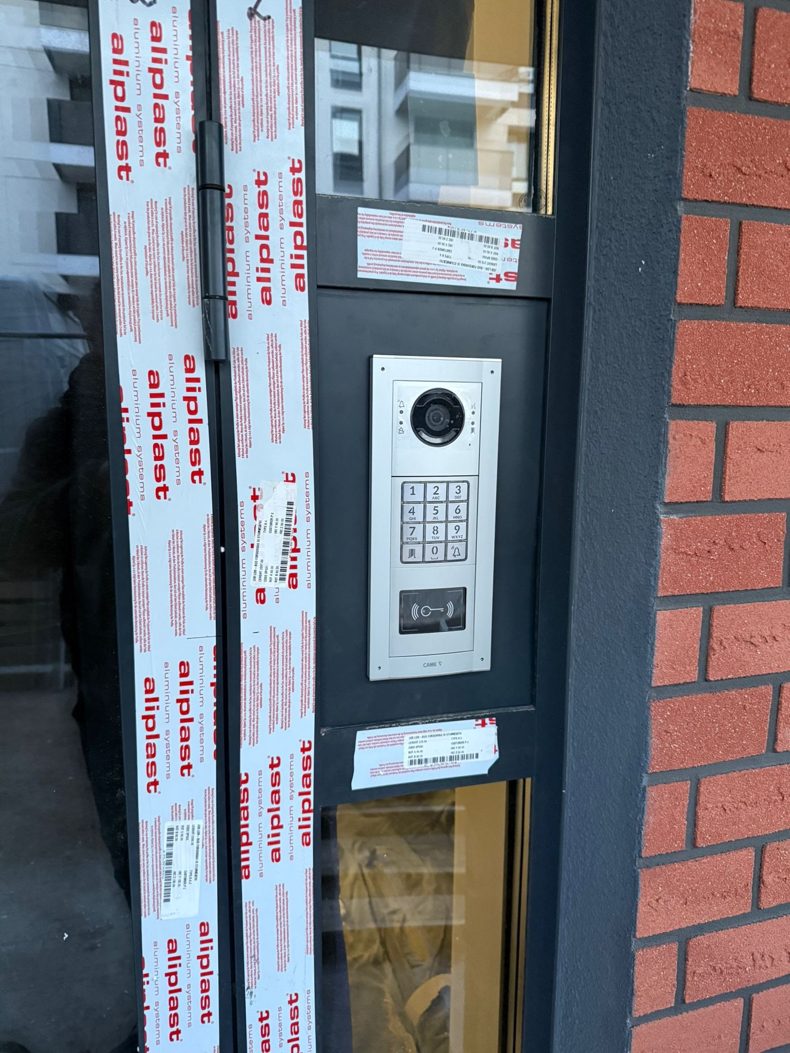Zamontowany wideodomofon CAME z klawiaturą numeryczną na tle ceglanej ściany i okna z folią ochronną Aluplast, widok z bliska na elewacji budynku.