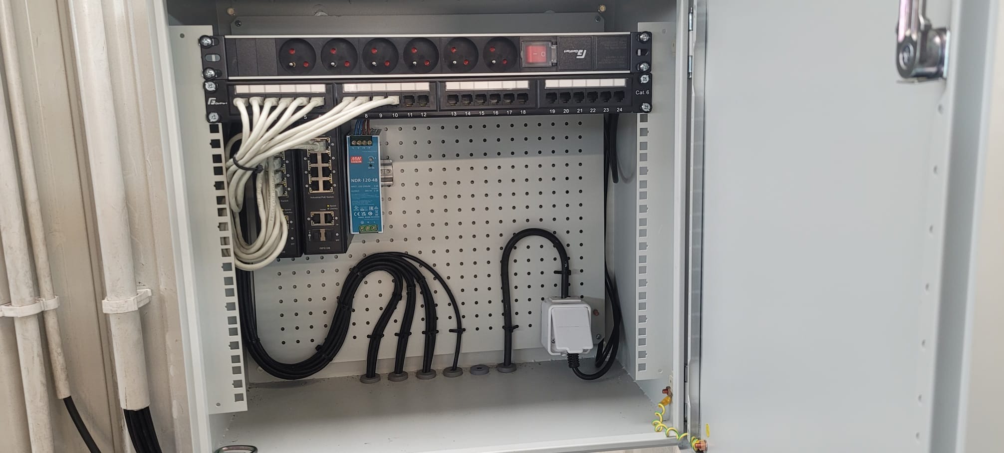 Otwarta szafa elektryczna z listwą zasilającą, patch panelem, switchem i okablowaniem. Widoczne gniazda, kable Ethernet, zasilacz i uziemienie.
