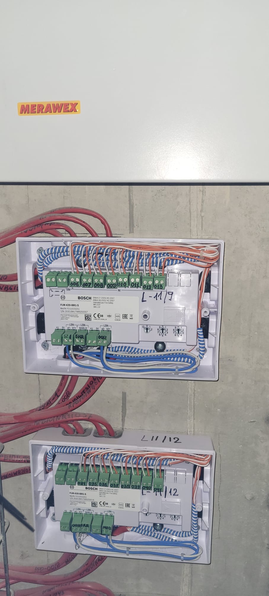 Dwa białe moduły Bosch FLM-420-IBR1-S z podłączonymi przewodami w kolorach czerwonym, niebieskim i pomarańczowym, zamontowane na szarej ścianie. Widoczne oznaczenia L-11/9 i L-11/12.