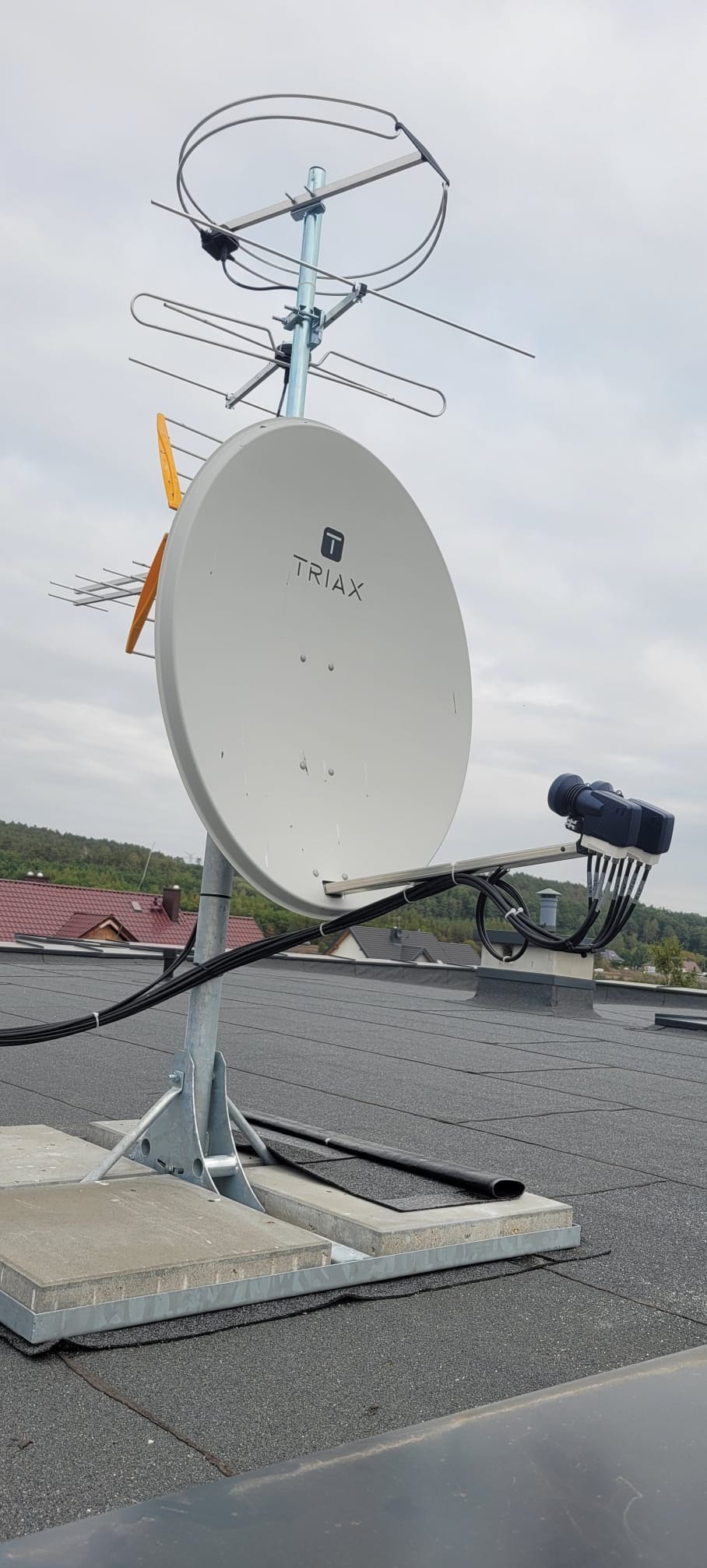 Antena satelitarna Triax i antena naziemna na dachu budynku z widokiem na zalesione wzgórza w tle, instalacja kabli.