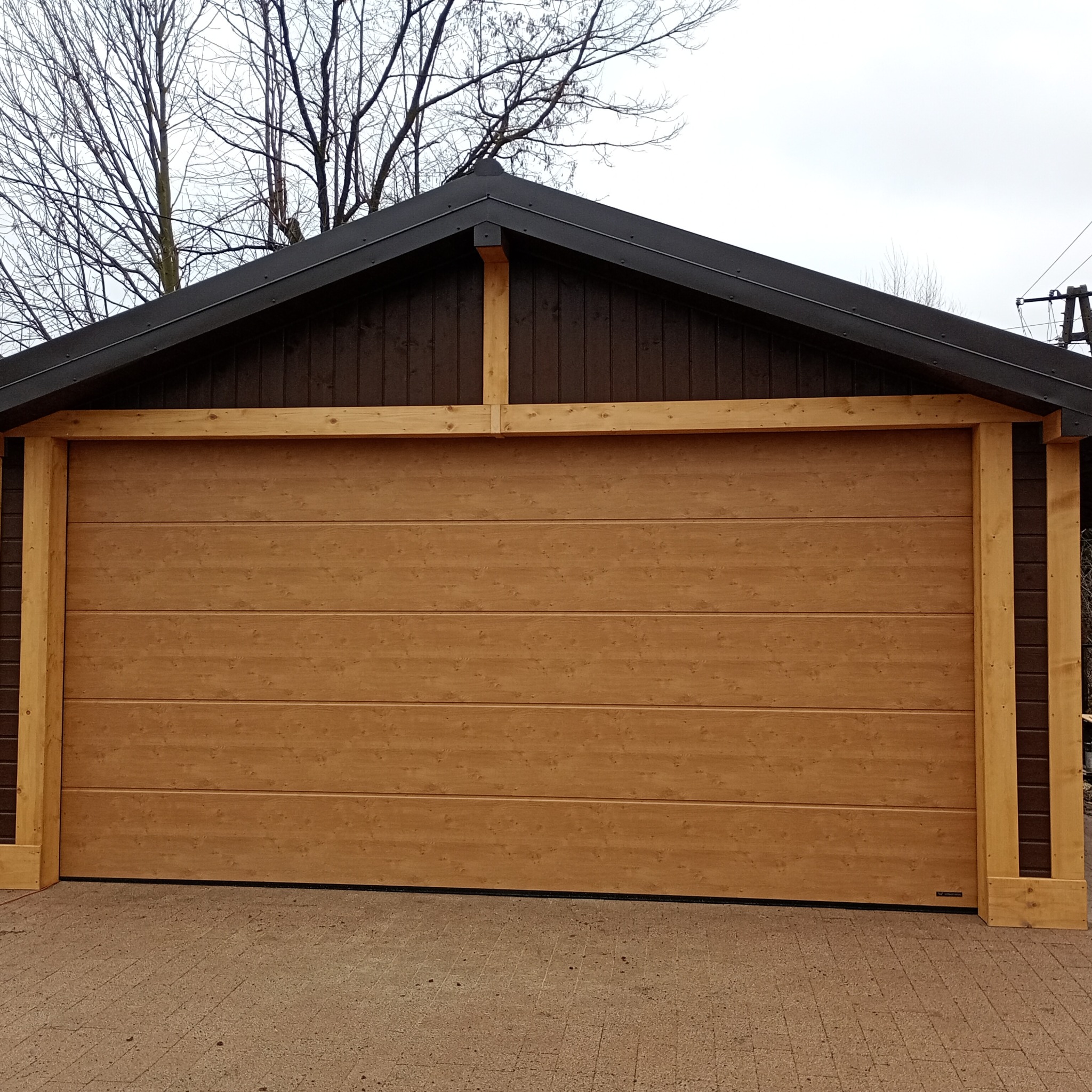Drewniana konstrukcja z bramą garażową o panelowej strukturze i jasnobrązowym kolorze, otoczona elementami z surowego drewna na tle pochmurnego nieba i drzew bez liści.