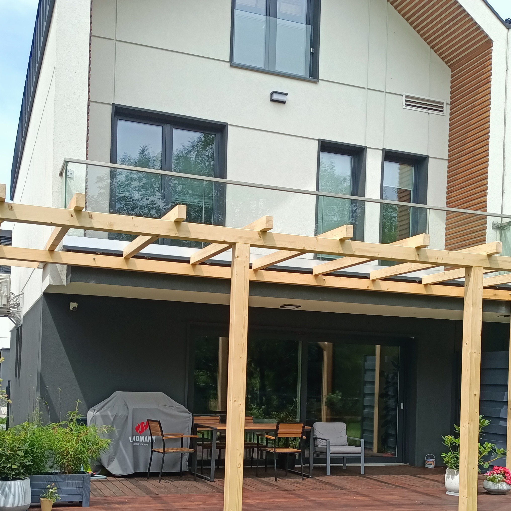 Nowoczesny dom z drewnianą konstrukcją zadaszenia tarasu, widoczne meble ogrodowe i osłona na grilla.