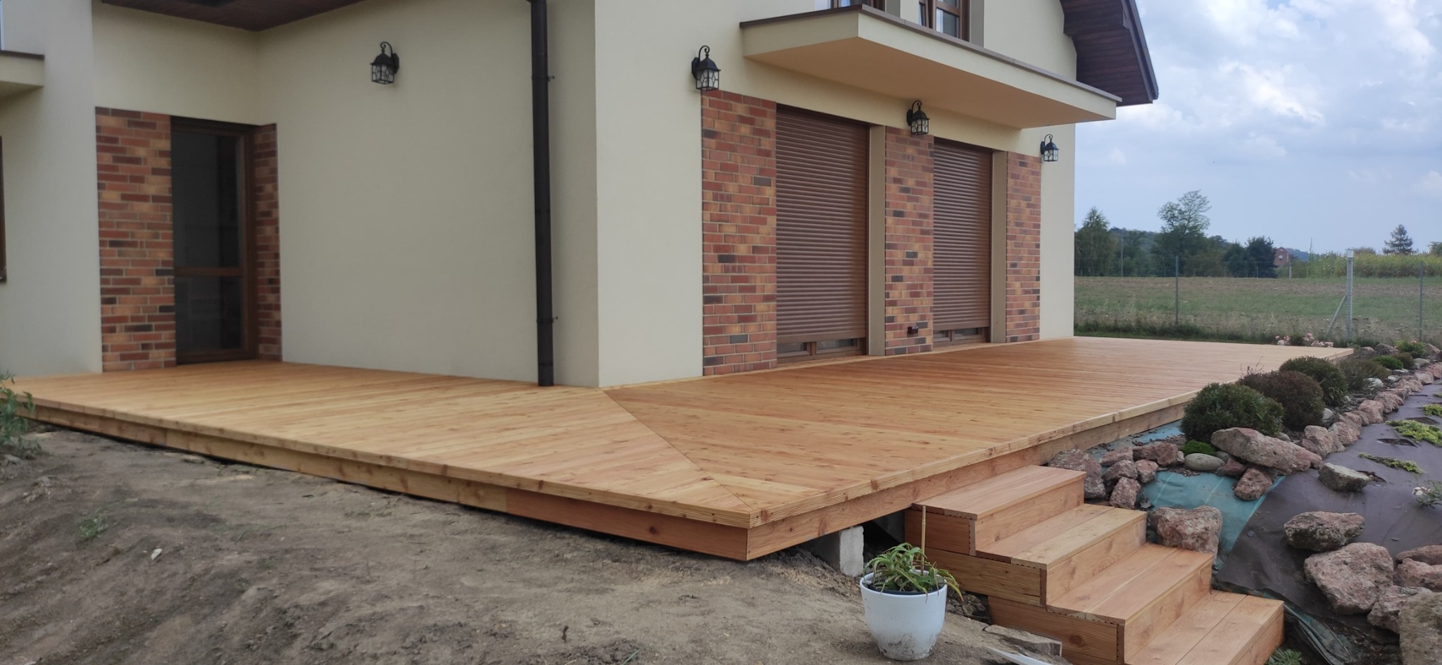 Przestronny taras z drewna przy domu z ceglaną elewacją, schody prowadzą do ogrodu. Jasne drewno, widoczne lampy na ścianie budynku.
