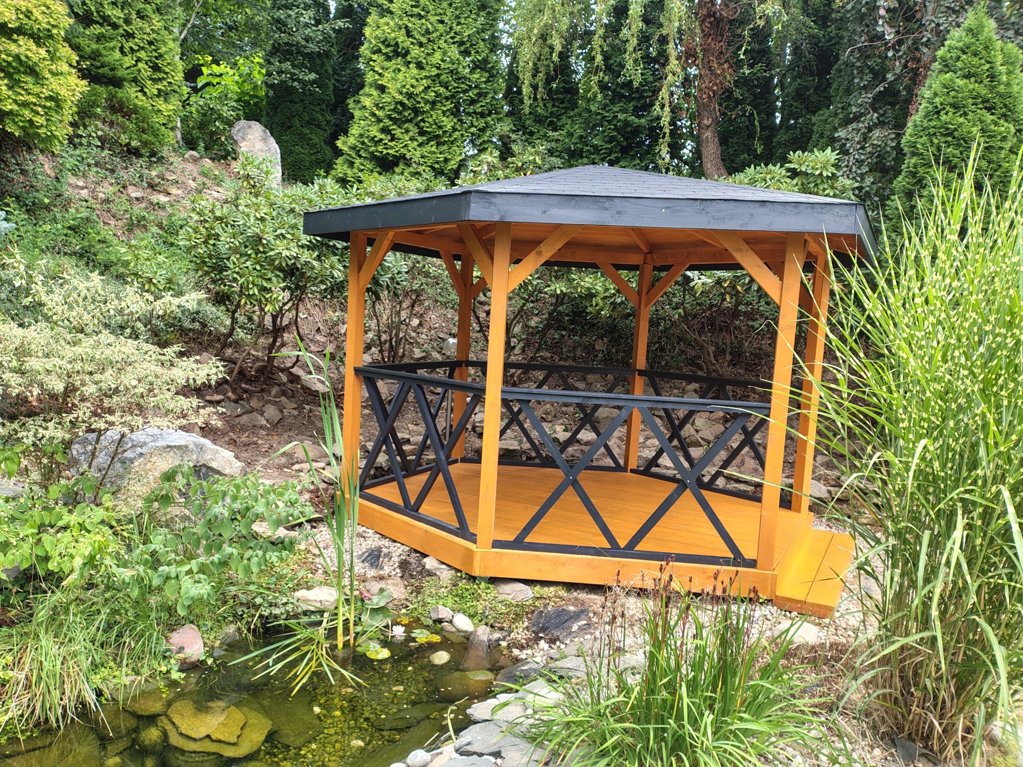 Ośmiokątna altana z drewnianą podłogą i czarnymi balustradami, otoczona zielenią i oczkiem wodnym, wkomponowana w naturalny krajobraz ogrodu.