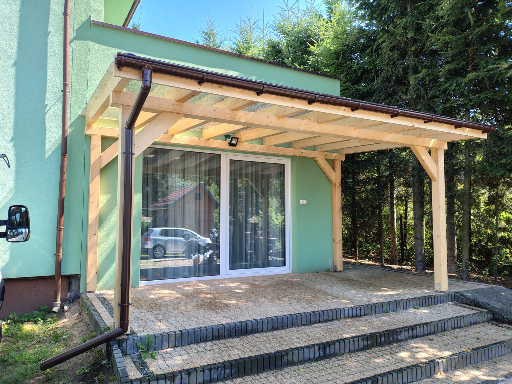 Zadaszenie tarasu z surowego drewna, wsparte na słupach, z brązowymi rynnami. W tle zielony budynek i drzewa. Widoczne schody z kostki brukowej.