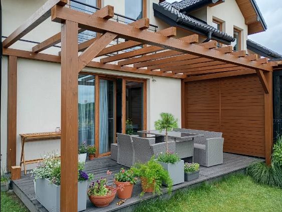 Drewniana pergola z meblami ogrodowymi na tarasie, z zabudową z desek w tle, kwiaty w donicach, widok na dom z czarnym dachem.