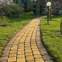 ChłopakiDobrzeRobią - Ścieżka z kostki brukowej w ogrodzie prowadząca do drewnianej altany z lampą, trawnik po bokach, w tle kwitnąca magnolia.