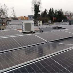 Widok z góry na instalację paneli słonecznych na płaskim dachu z widocznymi jednostkami zewnętrznymi klimatyzacji. Panele pokryte kroplami wody, w tle budynki mieszkalne i drzewa pod pochmurnym...