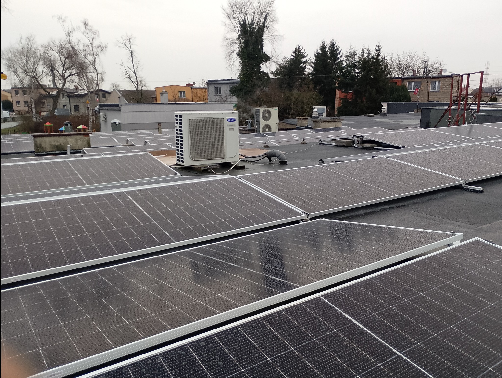 Widok z góry na instalację paneli słonecznych na płaskim dachu z widocznymi jednostkami zewnętrznymi klimatyzacji. Panele pokryte kroplami wody, w tle budynki mieszkalne i drzewa pod pochmurnym...