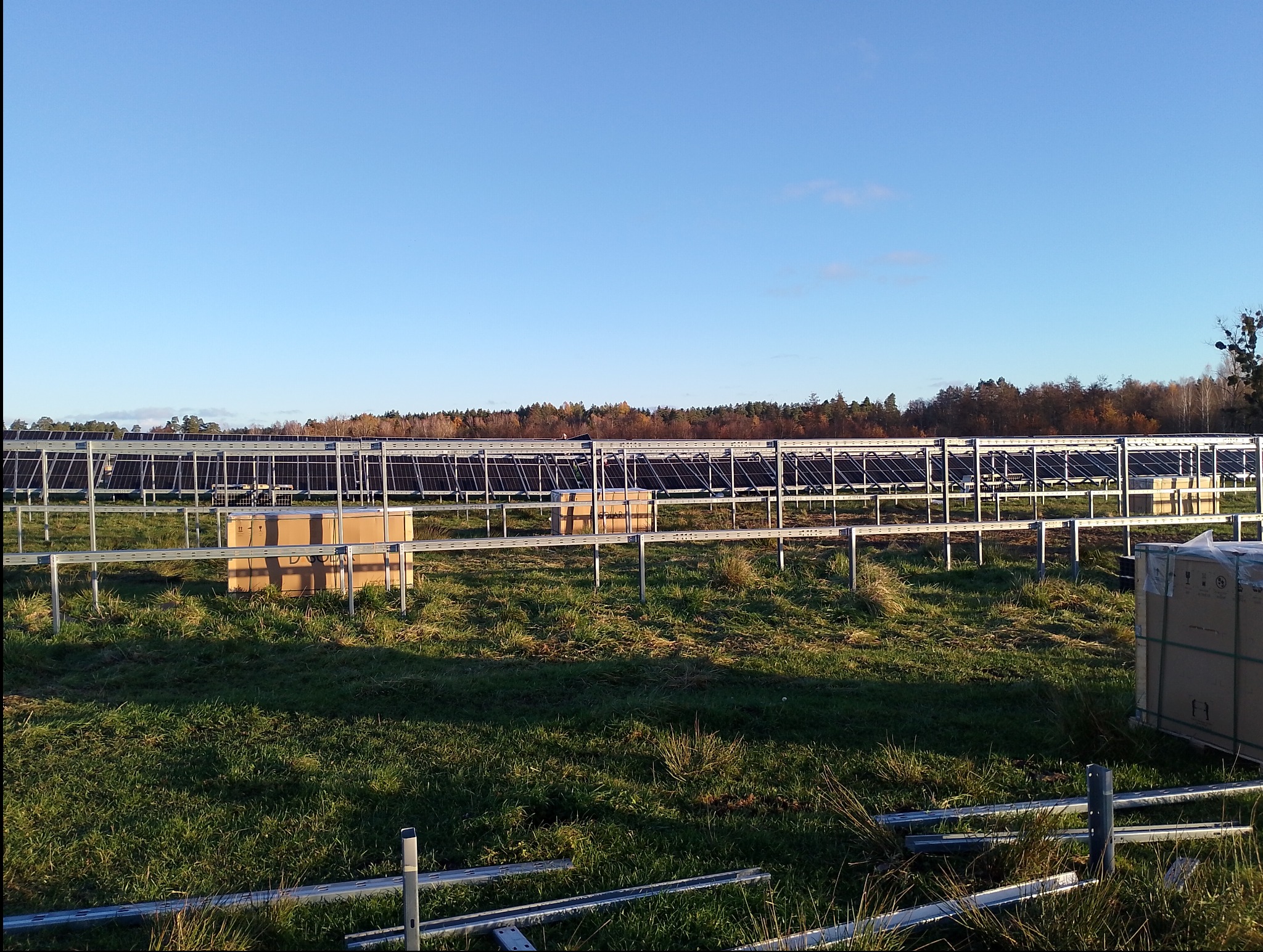 Rozległa instalacja fotowoltaiczna w trakcie montażu, widoczne rzędy konstrukcji wsporczych i panele słoneczne w oddali, na pierwszym planie kartonowe opakowania i elementy montażowe na zarośniętym...