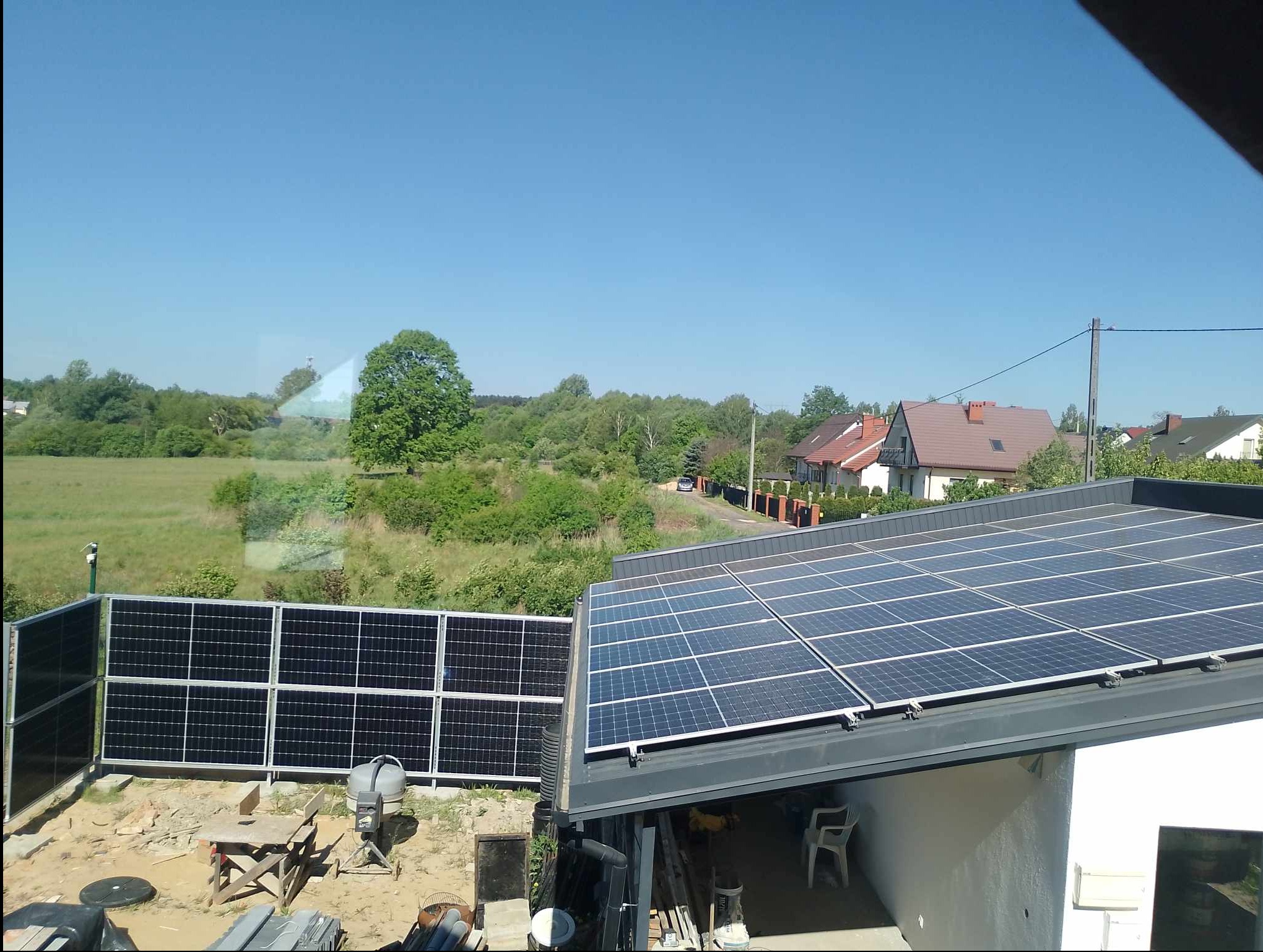 Panele słoneczne na dachu budynku, widok z góry na instalację i okolicę z zielenią oraz domami w tle.