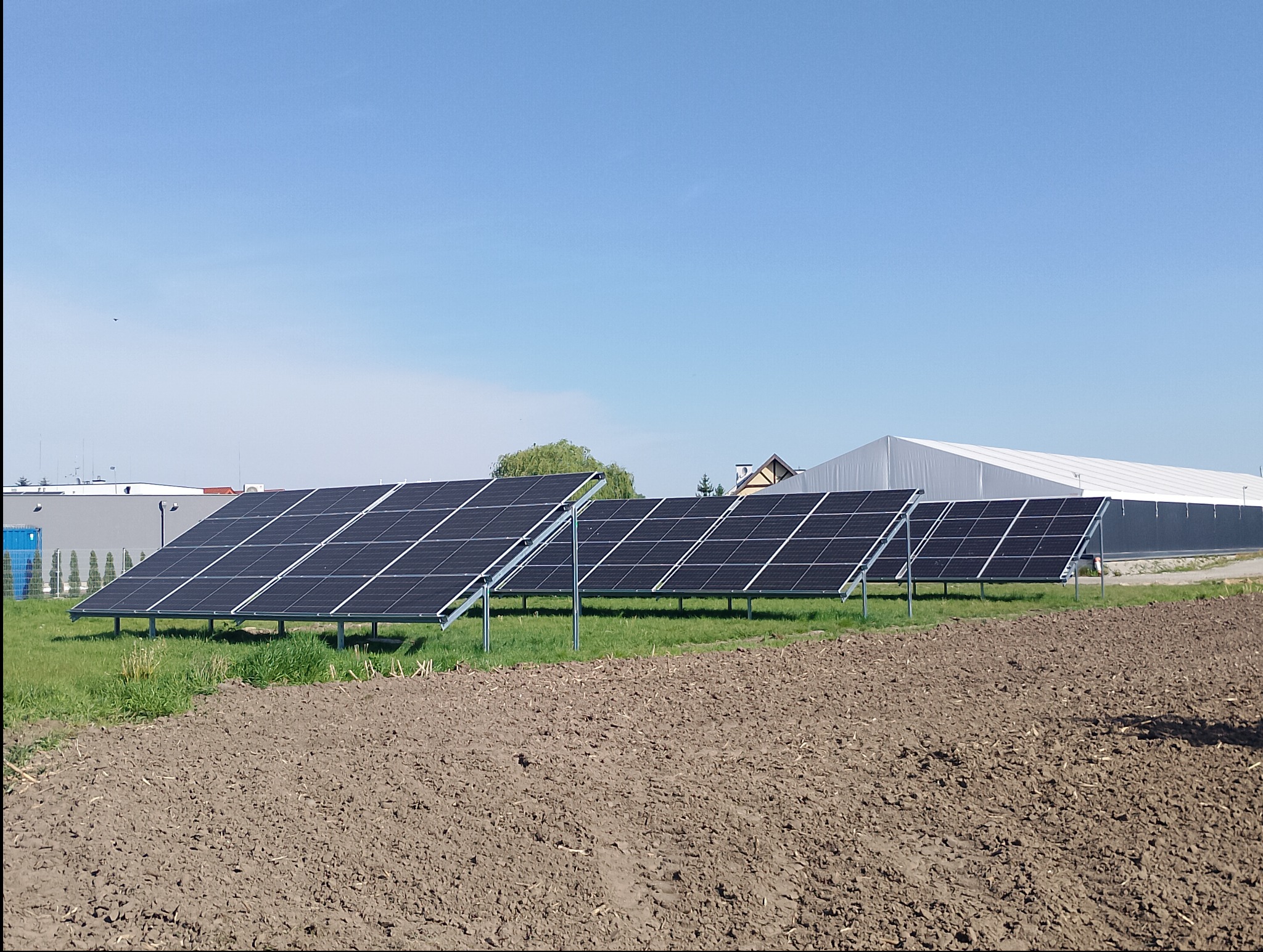 Rząd paneli słonecznych na metalowych stelażach, zainstalowanych na polu z przygotowaną glebą, w tle budynki przemysłowe i magazyn z białym dachem.