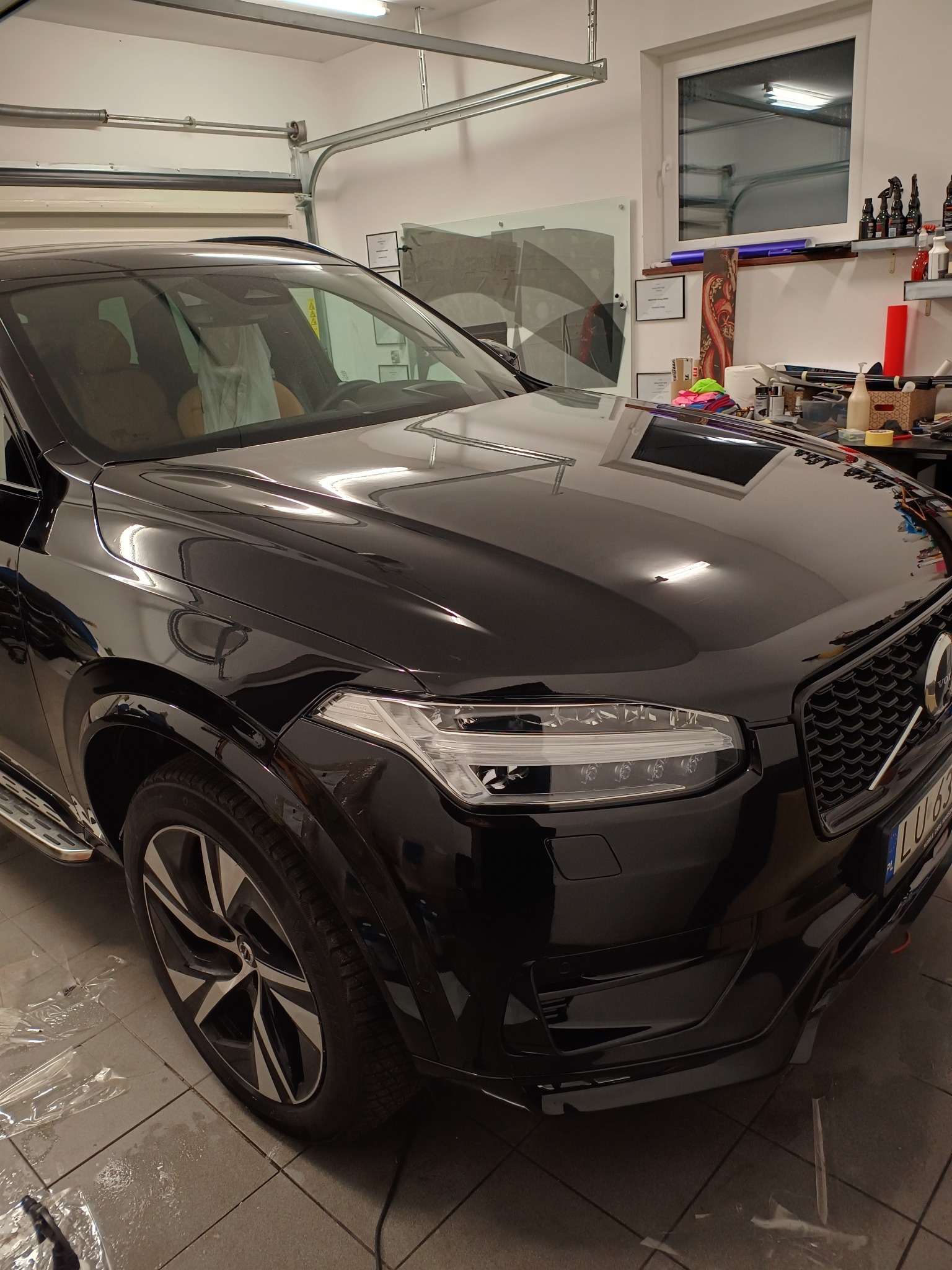 Czarny SUV Volvo XC90 po zabezpieczeniu folią ochronną, widoczne fragmenty folii na podłodze garażu, odbicie światła w lakierze, detale reflektorów i grilla.