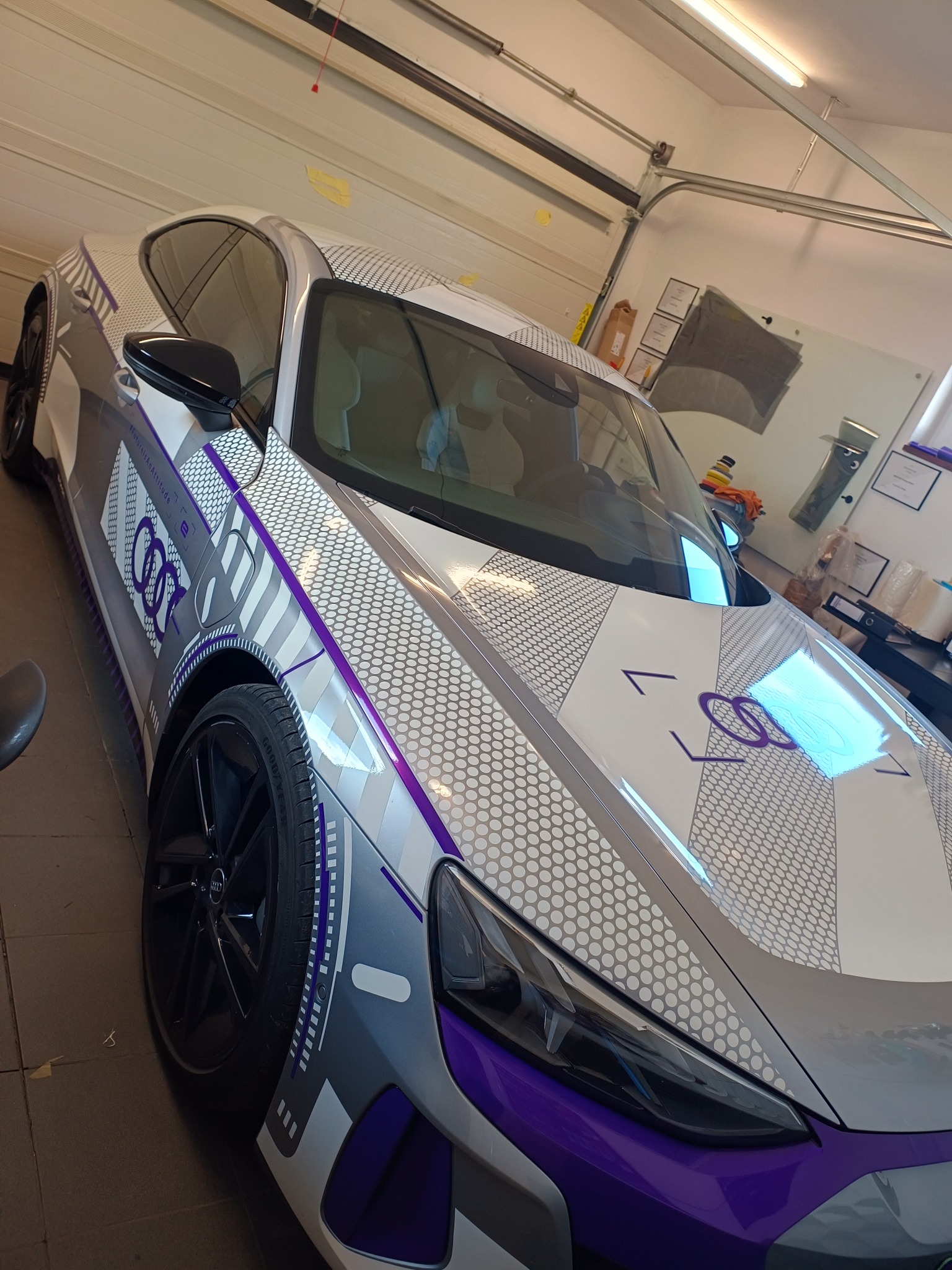 Audi E-Tron GT ICE RACE po oklejeniu, z widocznymi detalami grafiki w kolorach białym, szarym i fioletowym, w warsztacie.