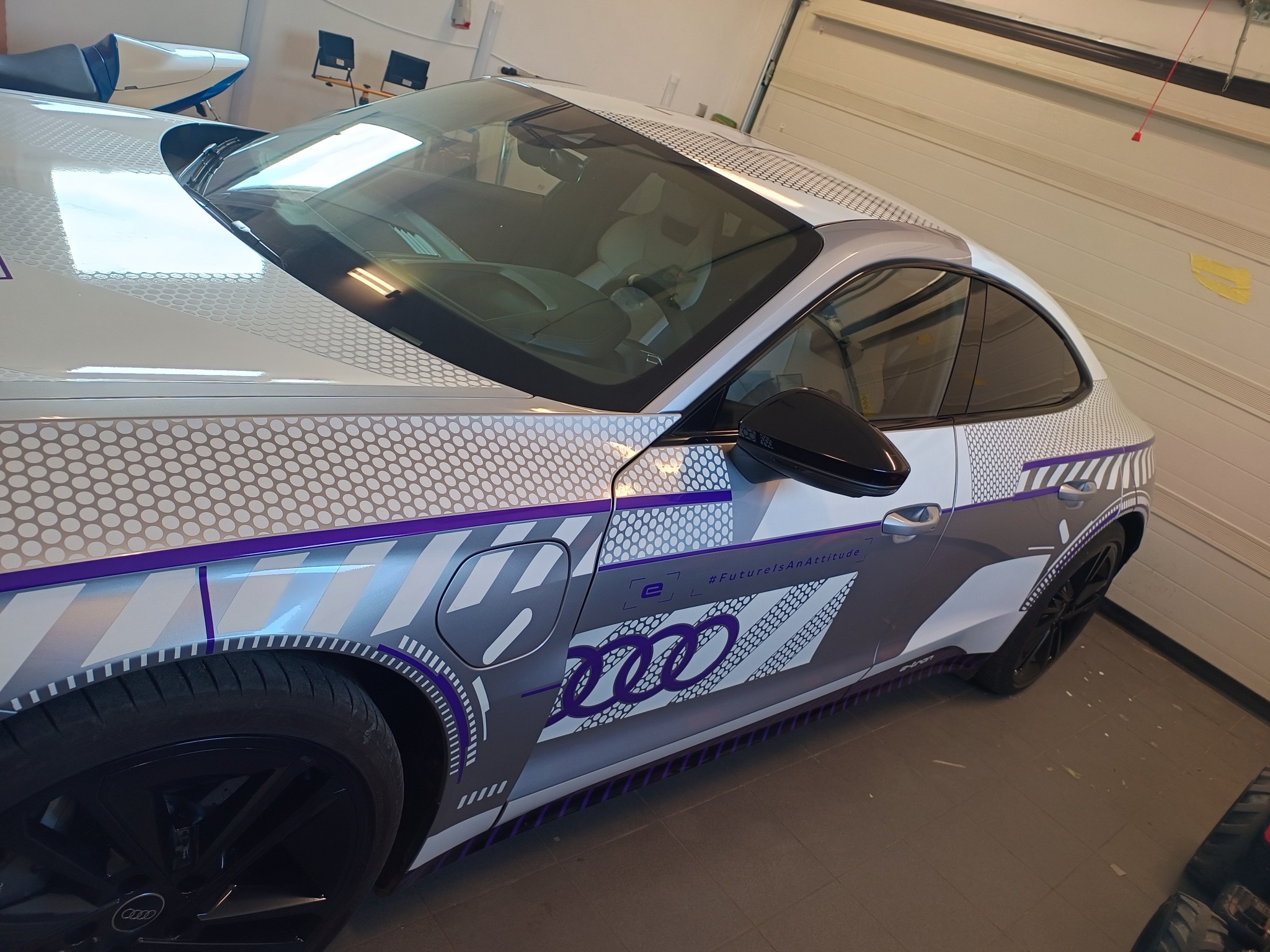 Białe Audi E-Tron GT (ICE RACE) z charakterystycznym oklejeniem fabrycznym w szaro-biało-fioletowe wzory, widoczne z boku, zaparkowane w garażu.