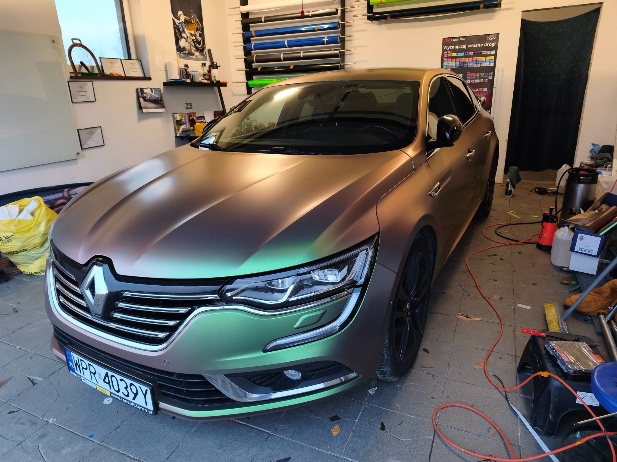 Renault po zmianie koloru folią, z efektem kameleona, w warsztacie. Widoczne narzędzia i wzorniki folii na ścianie. Unikalny odcień i połysk.