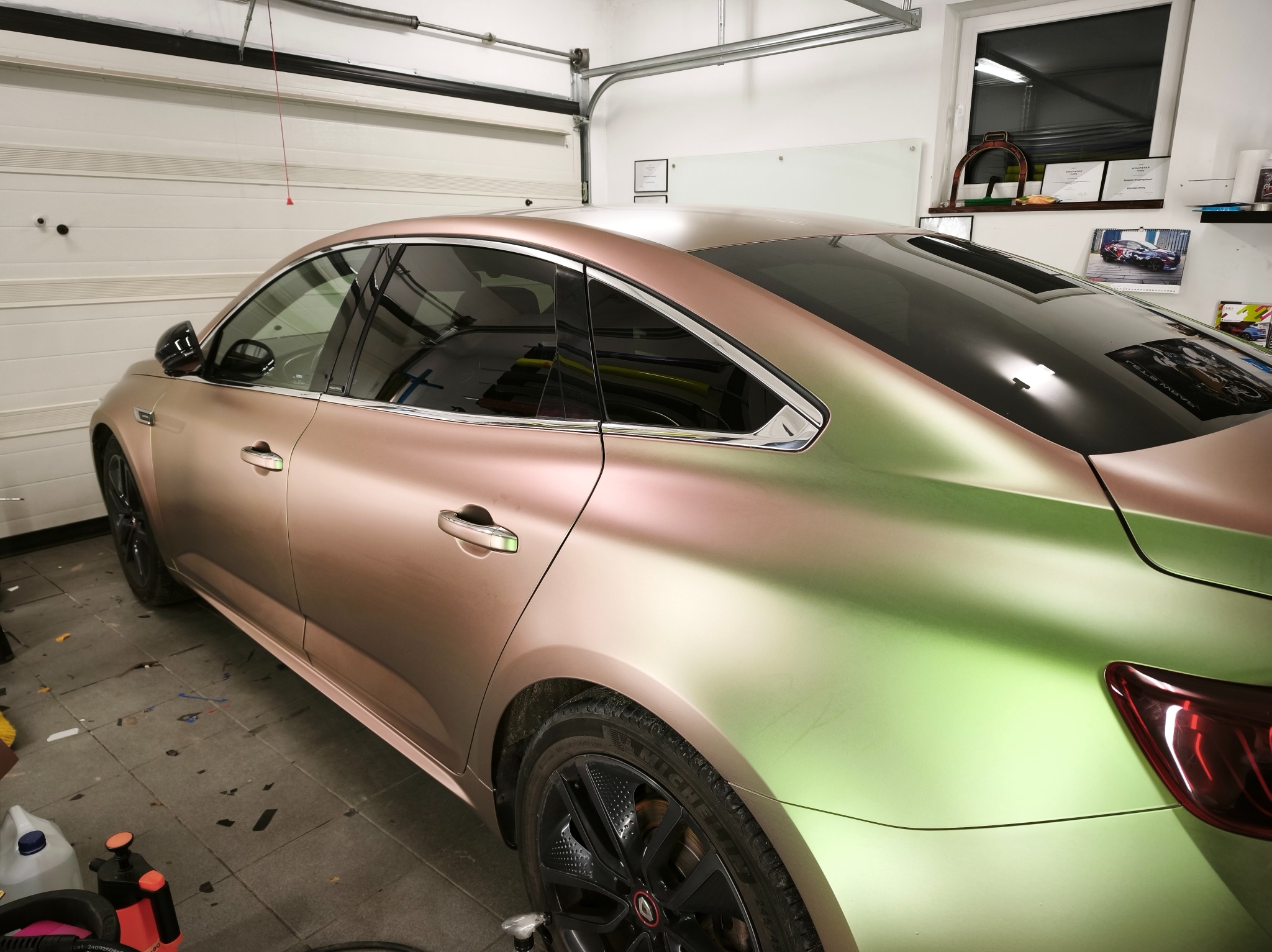 Auto po oklejeniu folią w odcieniach różu i zieleni, widoczne w garażu z podniesioną bramą. Na podłodze resztki folii i narzędzia.