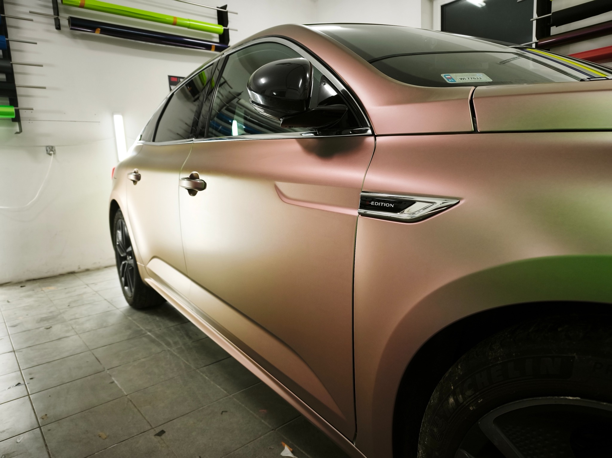 Auto w matowej folii w odcieniach różu i zieleni, z widocznym logo 'Edition' na błotniku, zaparkowane w warsztacie z rolkami folii na ścianie.