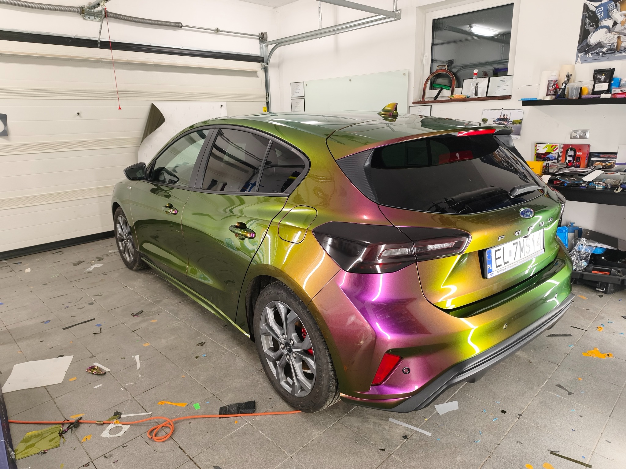 Ford Focus po oklejeniu folią zmieniającą kolor, od zieleni po fiolet, w garażu z widocznymi narzędziami i resztkami folii na podłodze. Efektowna metamorfoza.
