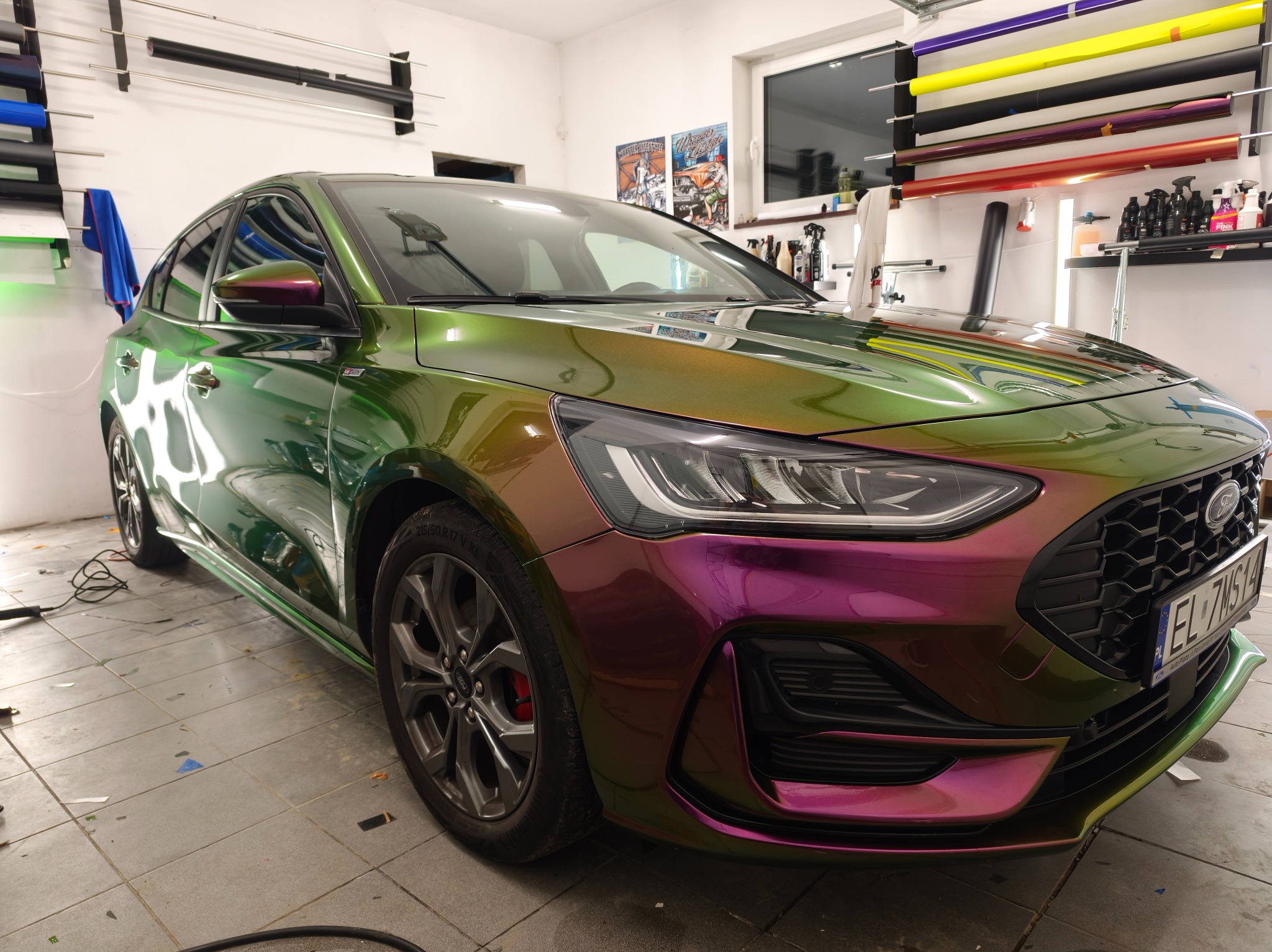 Ford Focus po oklejeniu folią zmieniającą kolor, od zieleni po fiolet, w warsztacie z rolkami folii na ścianie. Widoczne detale: felgi, reflektory i tablica rejestracyjna.