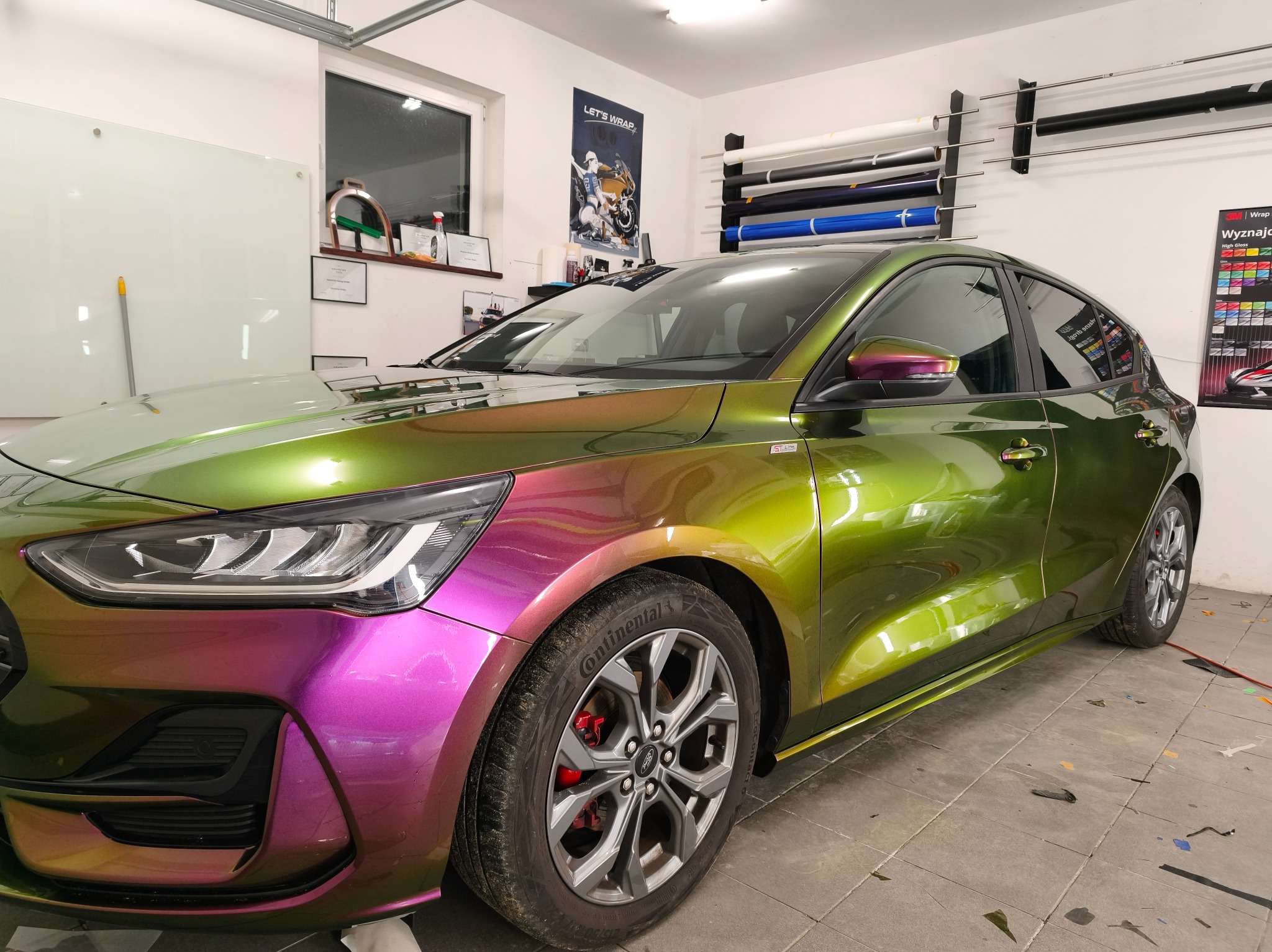 Ford Focus po oklejeniu folią zmieniającą kolor, od fioletu do zieleni, w warsztacie. Widoczne rolki folii i plakat reklamowy. Felgi aluminiowe i opony Continental.