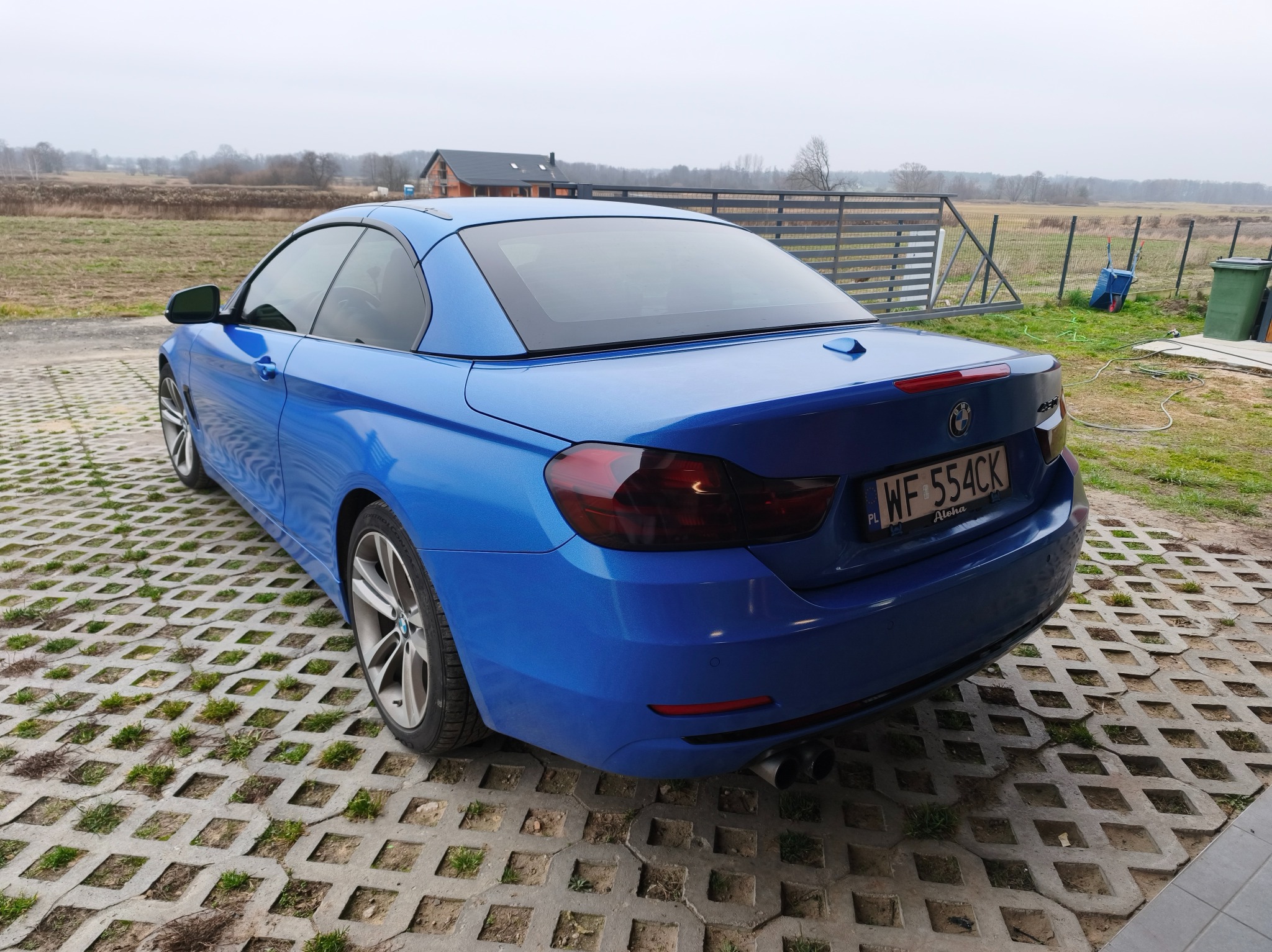 Błękitne BMW 4 Cabrio na ażurowym podłożu, widok z tyłu. Auto z przyciemnionymi szybami, na tle krajobrazu z domem w budowie i polami.