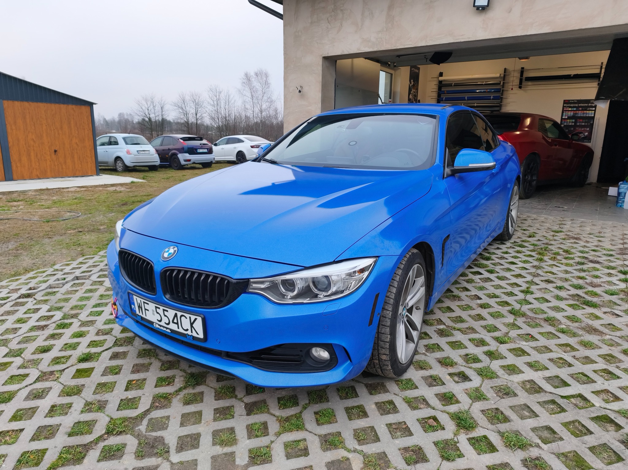 Błękitne BMW serii 4 po oklejeniu folią, zaparkowane na ażurowym podjeździe. W tle budynek warsztatu z widocznym czerwonym samochodem i regałem z foliami.