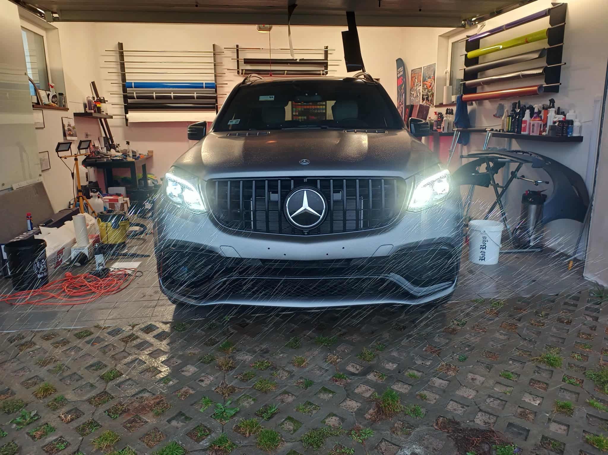Srebrny Mercedes po aplikacji folii ochronnej, widoczne rolki folii i narzędzia w tle garażu. Oświetlenie podkreśla detale karoserii.