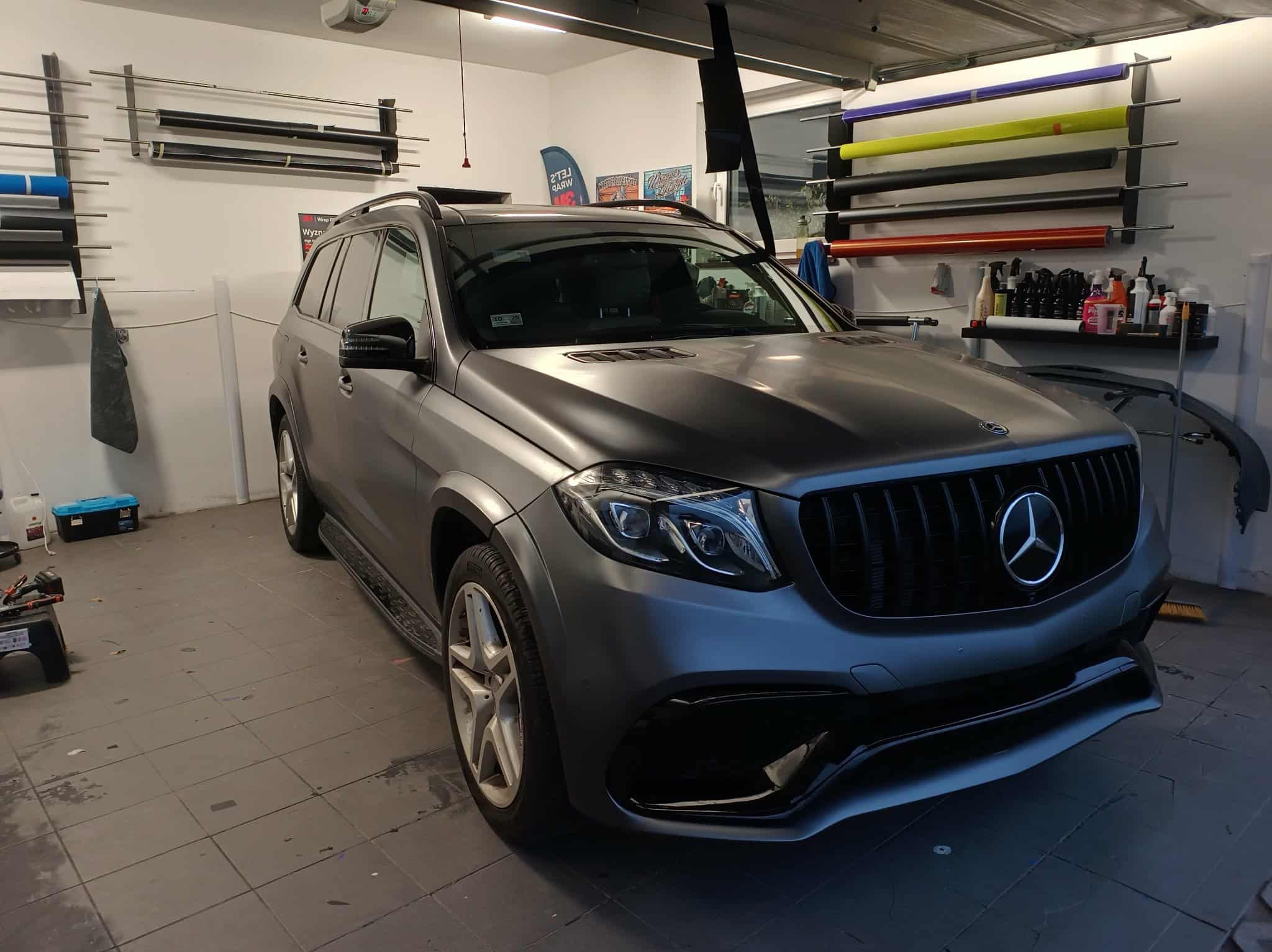 Matowy Mercedes-Benz GLS po oklejeniu folią w warsztacie, widoczne rolki folii na ścianie. Auto z czarnym grillem i srebrnymi felgami, oświetlenie warsztatowe.