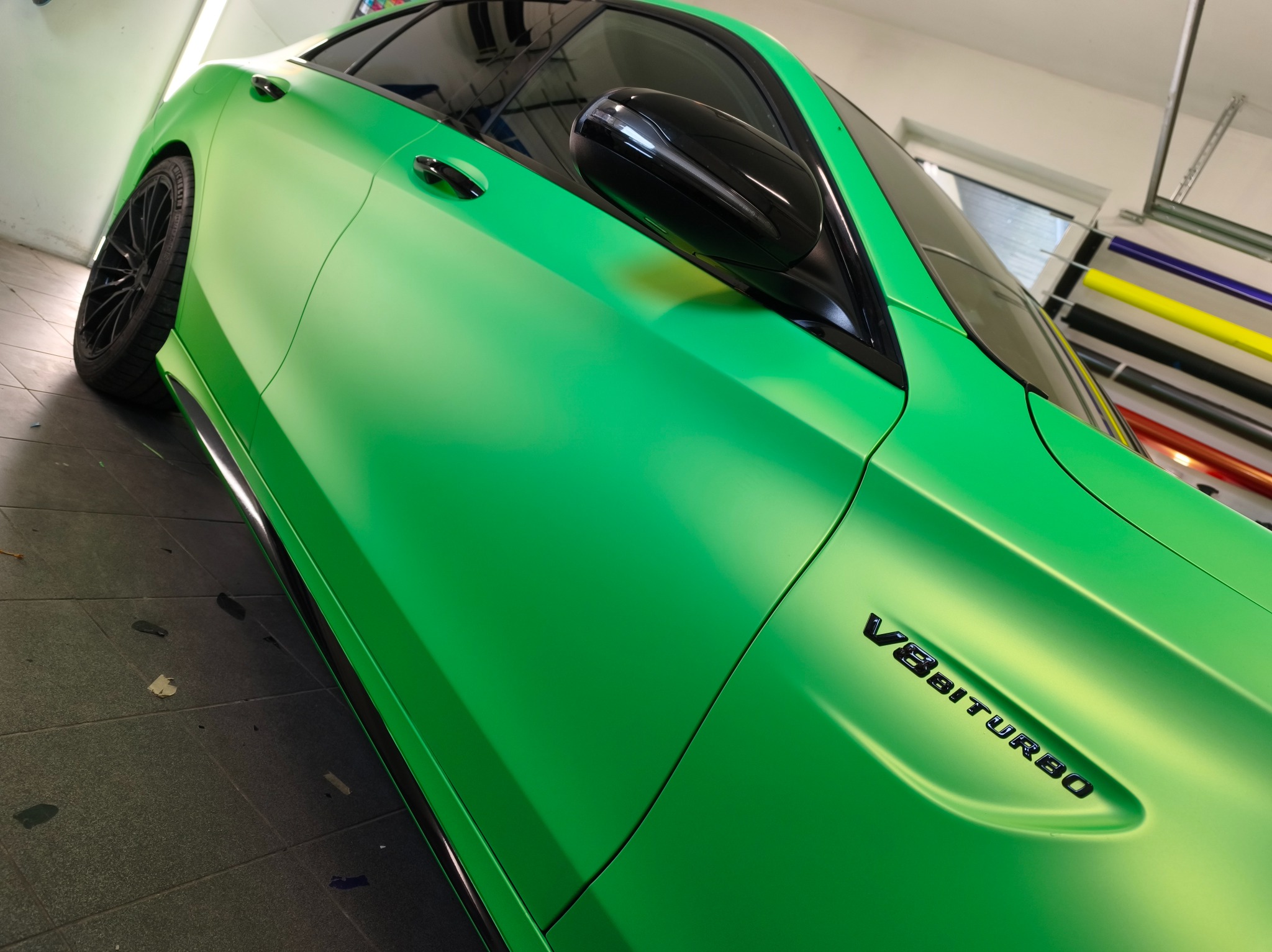 Zielony, matowy Mercedes-AMG GT po oklejeniu folią w Kąck. Widoczny napis V8 BITURBO. W tle rolki z foliami do oklejania.