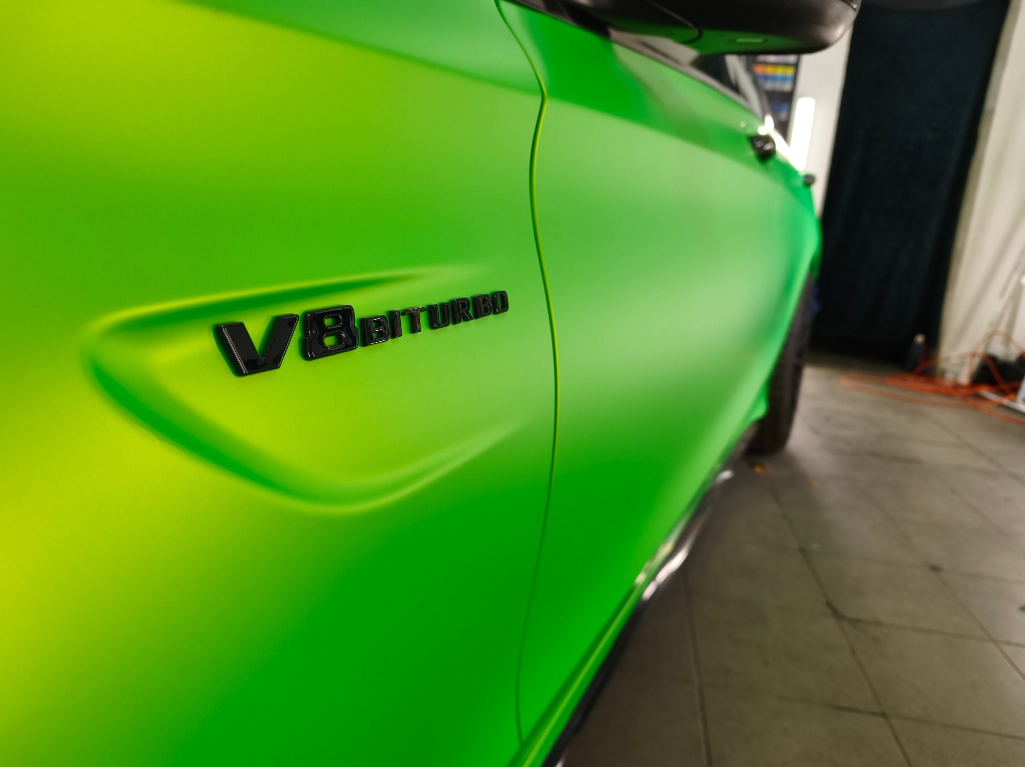 Jaskrawozielony, matowy lakier na boku auta z czarnym napisem 'V8 BITURBO'. Widoczne detale karoserii i oświetlenie warsztatowe.
