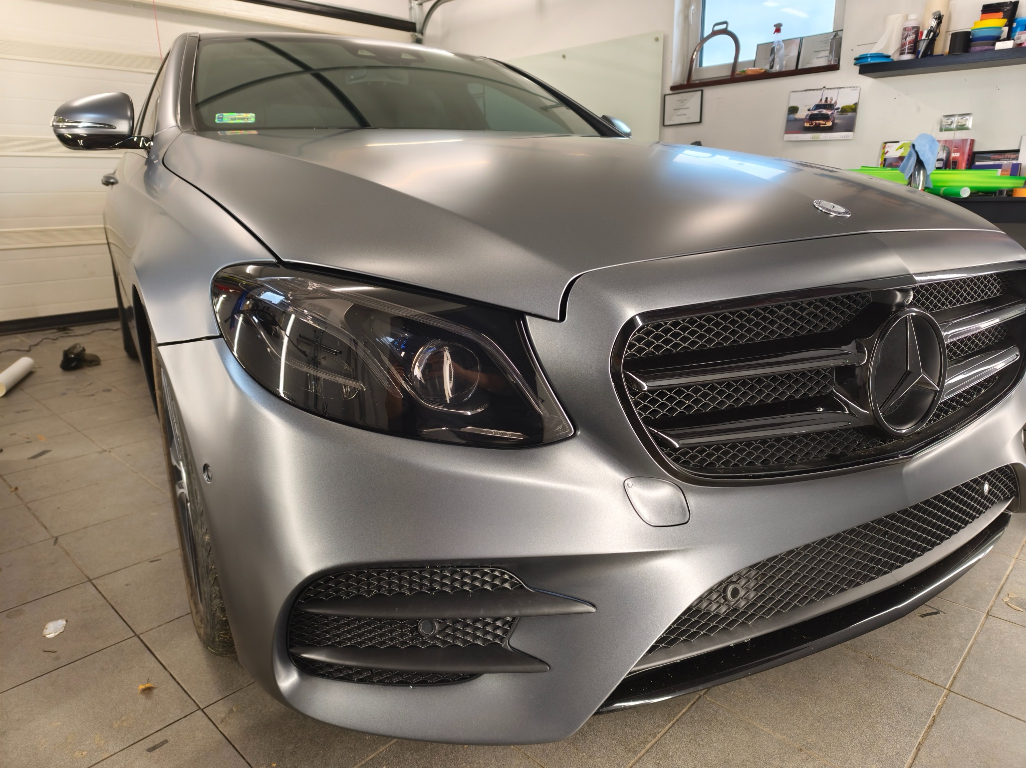 Matowy, srebrny Mercedes klasy E po zmianie koloru folią, widoczny z przodu, z detalami grilla i reflektorów. W tle warsztat.