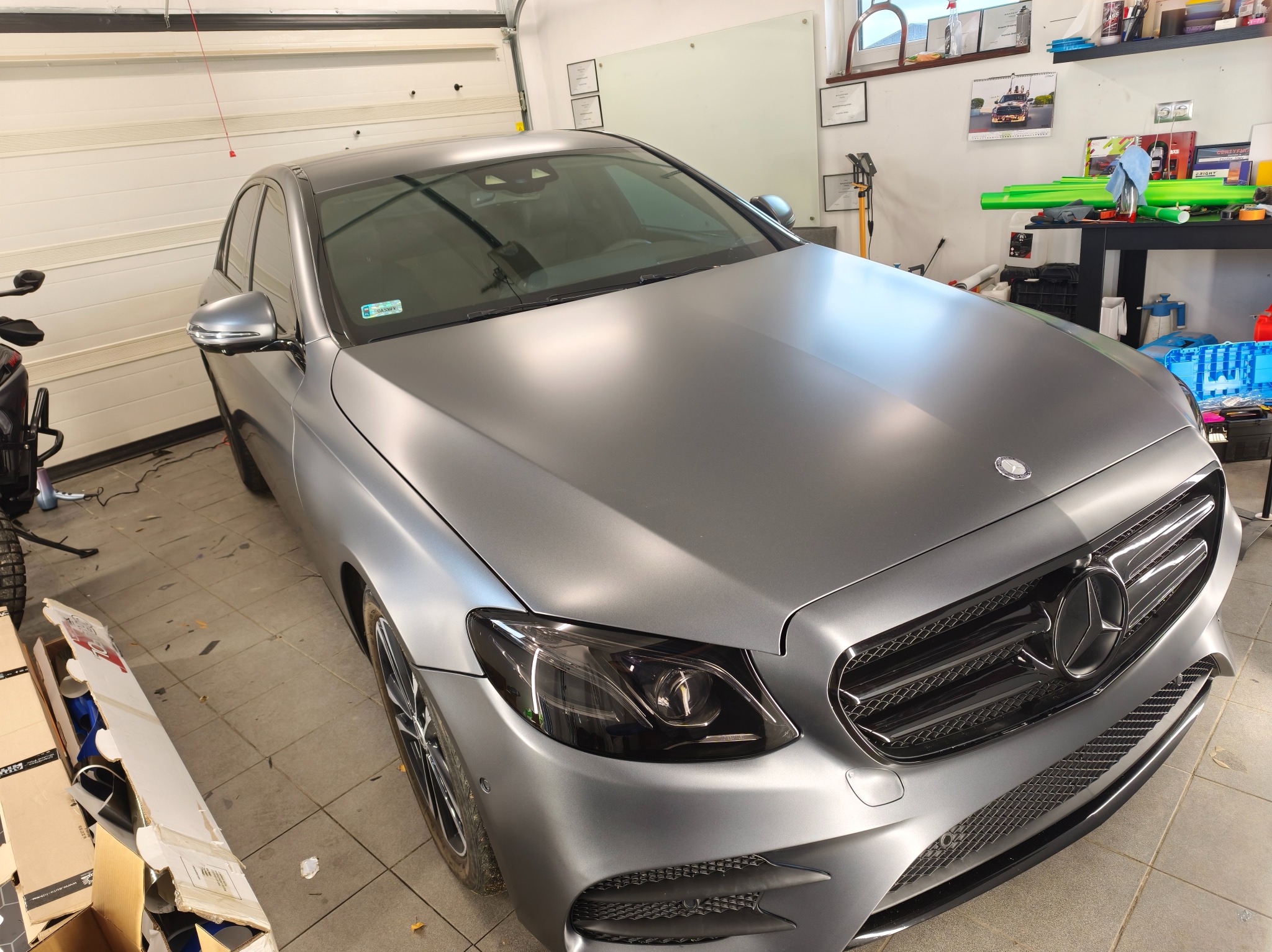 Szary Mercedes E-Klasa po oklejeniu matową folią, widoczny w garażu. Detal: przyciemnione reflektory i chromowana atrapa chłodnicy. Na ścianie certyfikaty i kalendarz.