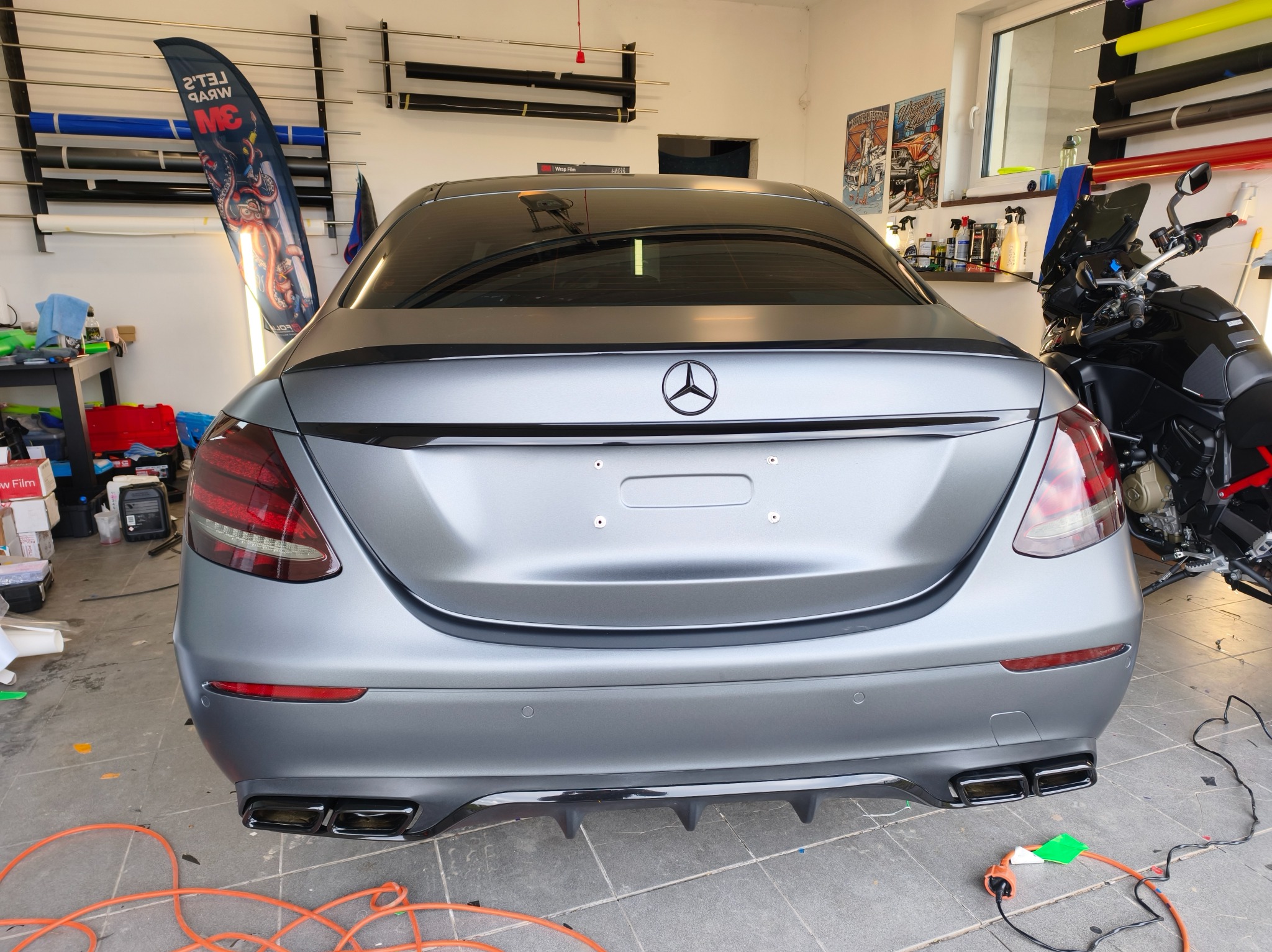 Szary Mercedes po oklejeniu folią matową, widok z tyłu. W tle narzędzia i materiały do oklejania, obok zaparkowany motocykl. Detal: czarne logo Mercedesa.