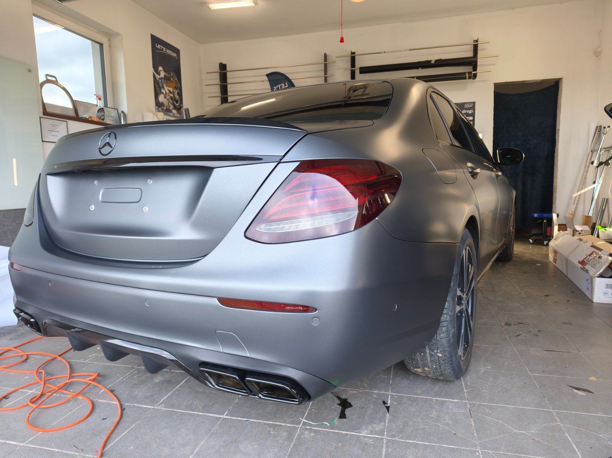 Matowy, szary Mercedes po oklejeniu folią, widoczny z tyłu w warsztacie. Detale: czarny dyfuzor, poczwórne końcówki wydechu, lampy. Pomarańczowy kabel na podłodze.