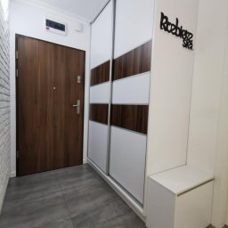Unikalna Forma Piotr Sulik - Przedpokój z dużą szafą przesuwną z białymi i drewnopodobnymi panelami, brązowe drzwi wejściowe, szara podłoga i biała szafka siedzisko