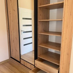 Unikalna Forma Piotr Sulik - Drewniana szafa z przesuwnymi drzwiami z lustrem, otwartymi półkami i wysuniętymi szufladami, widoczna w odbiciu biała drzwi z czarnymi paskami.