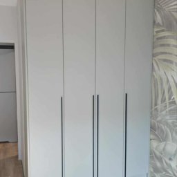 Unikalna Forma Piotr Sulik - Jasnoszara szafa z czterema drzwiami i długimi, czarnymi uchwytami, na tle ściany z motywem liści. Podłoga z paneli imitujących drewno. Widoczna żarówka bez klosza.