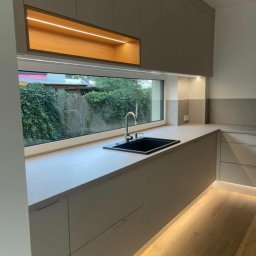 Unikalna Forma Piotr Sulik - Nowoczesna kuchnia z szarymi szafkami, czarnym zlewem i oknem z widokiem na zieleń. Podświetlenie LED pod szafkami i w górnej wnęce dodaje ciepła. Minimalistyczny design.