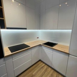 Unikalna Forma Piotr Sulik - Nowoczesna, narożna kuchnia z białymi, matowymi frontami i drewnianym blatem. Oświetlenie LED pod szafkami górnymi. Widoczna płyta indukcyjna i czarny zlew.