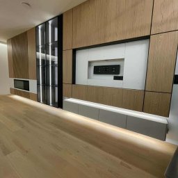 Unikalna Forma Piotr Sulik - Drewniana zabudowa ściany z szafkami, wnęką na TV i przeszkloną witryną z podświetleniem LED. Nowoczesny design, jasne drewno, minimalistyczny styl.