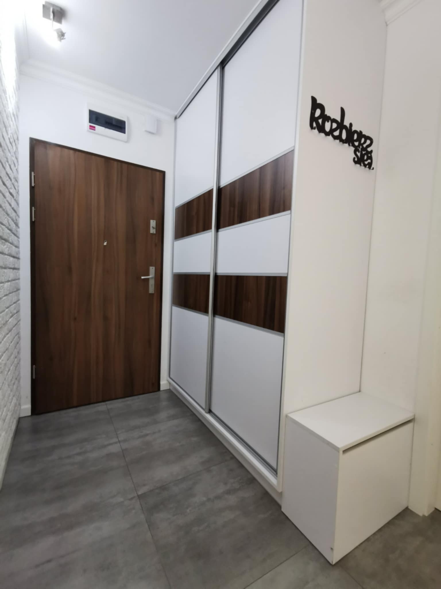 Przedpokój z dużą szafą przesuwną z białymi i drewnopodobnymi panelami, brązowe drzwi wejściowe, szara podłoga i biała szafka siedzisko